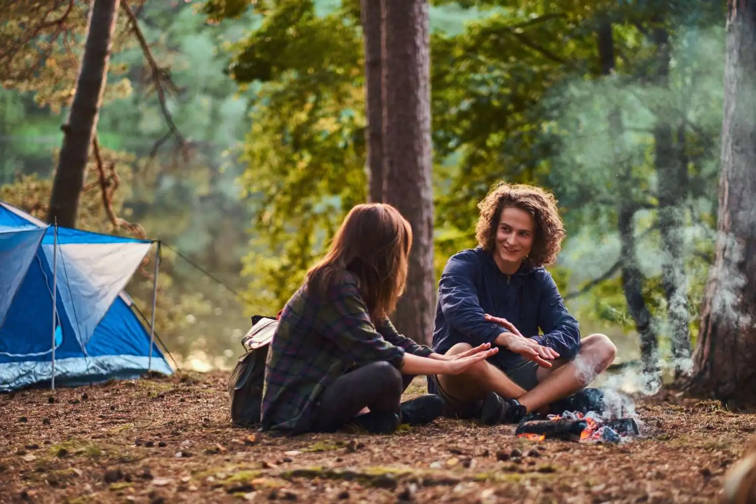 happy-young-couple-sitting-warming-near-campfire-camp-forest-travel-tourism-hike-concept